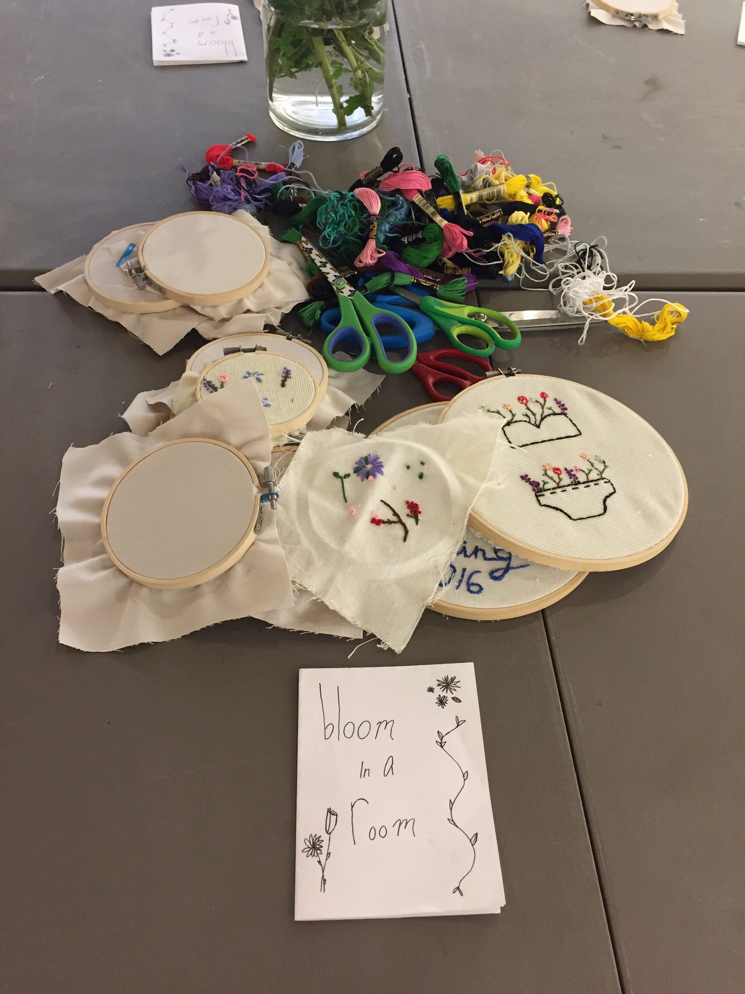 Various cross-stitch pieces on display with scissors, string and paper reading "bloom in a room".
