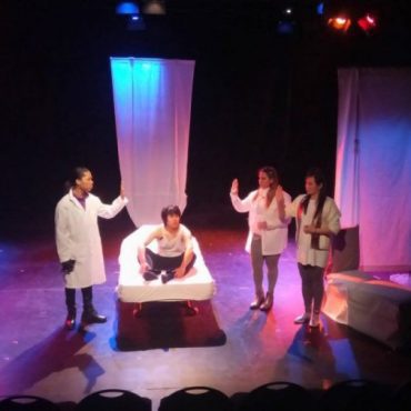 Four students on stage with white sheets hanging in background and man sitting on bed.