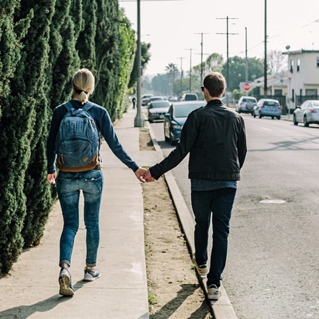 Woman with backpack on sidewalk and man on curb walking holding hands.