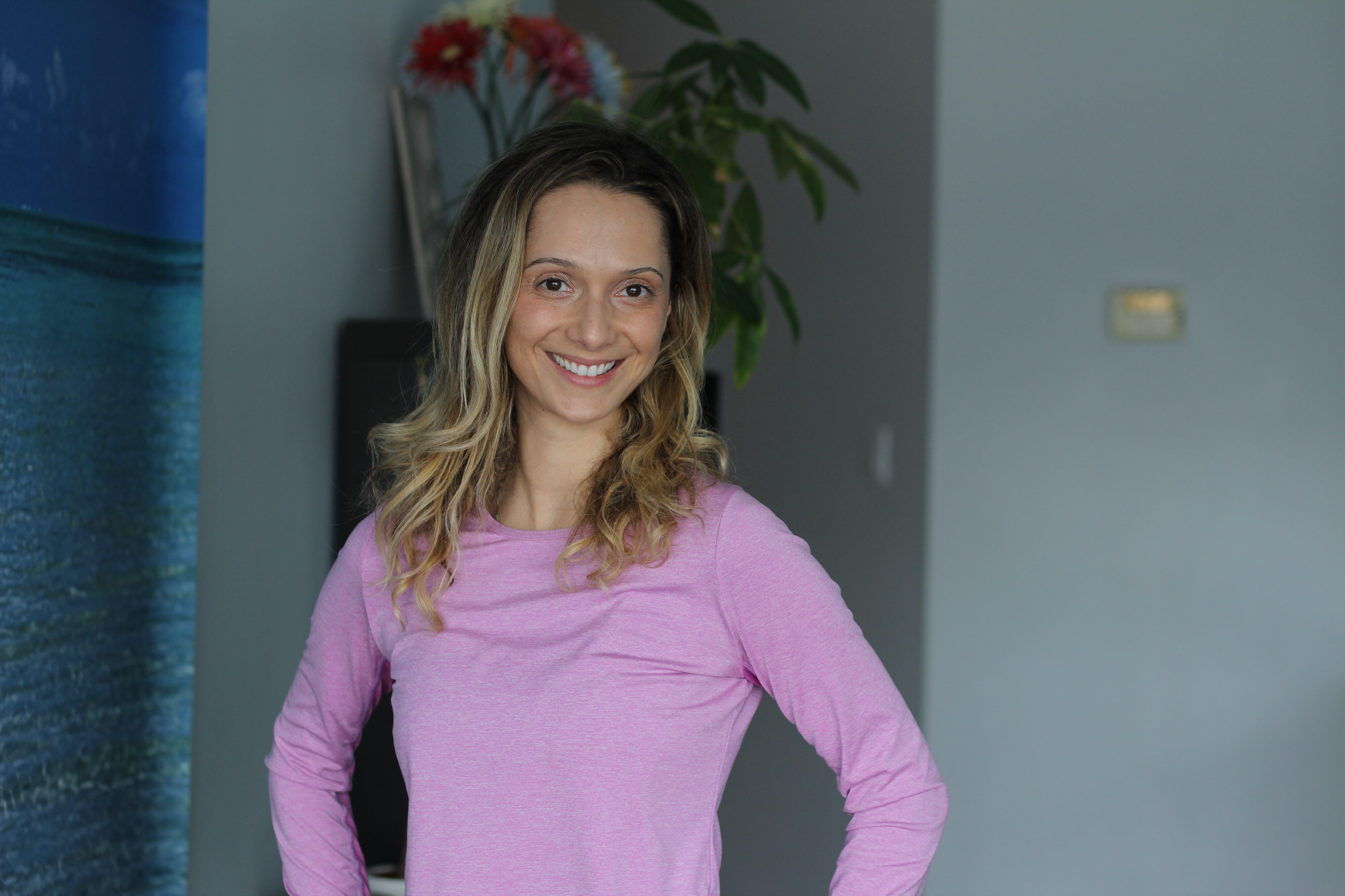 Portrait of yoga instructor Grabriela Nicole Doiu standing in pink yoga clothes in front of a shelf and plant.
