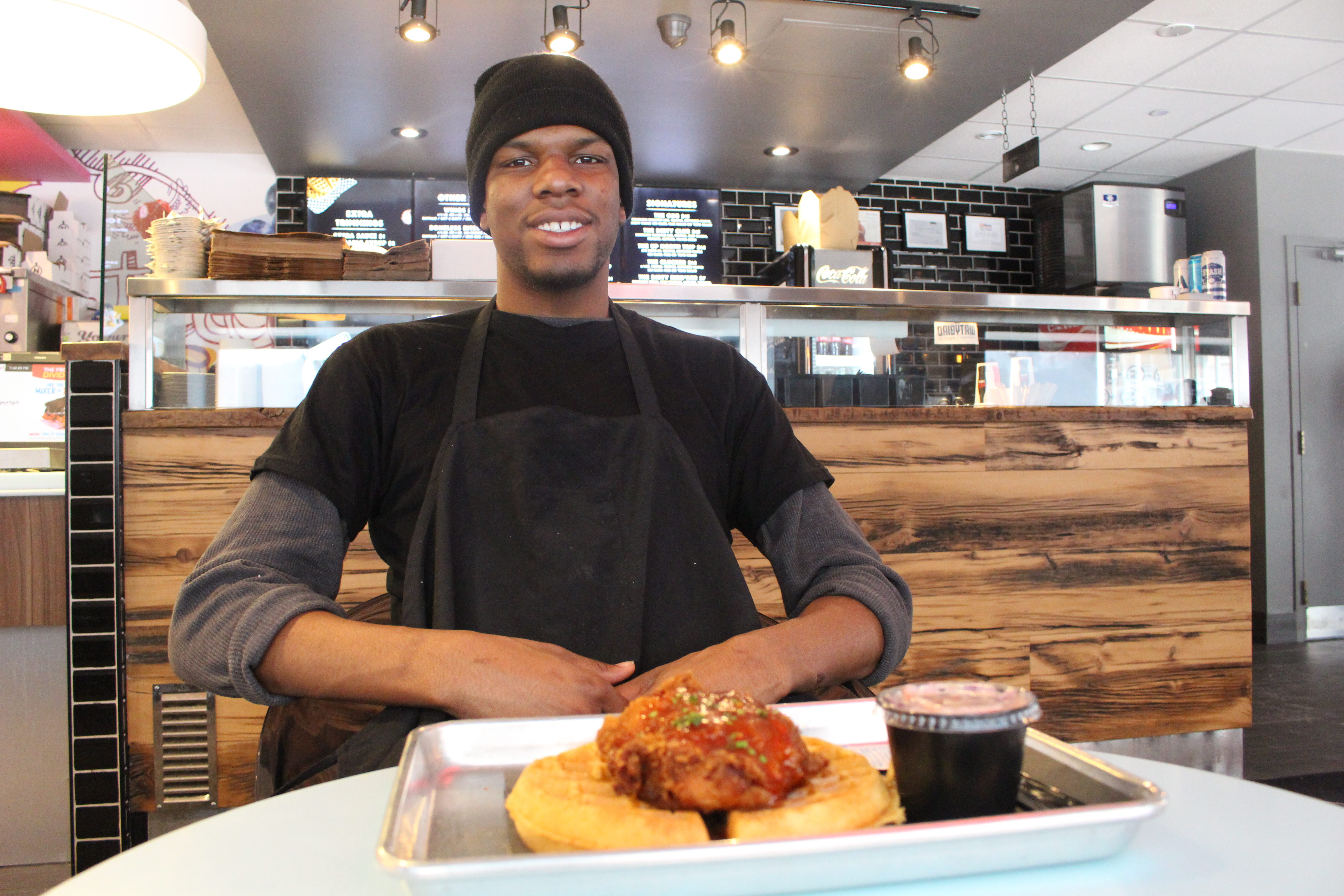 Cook Rashawan Farquason sitting in front of chicken and waffles with menu in background.