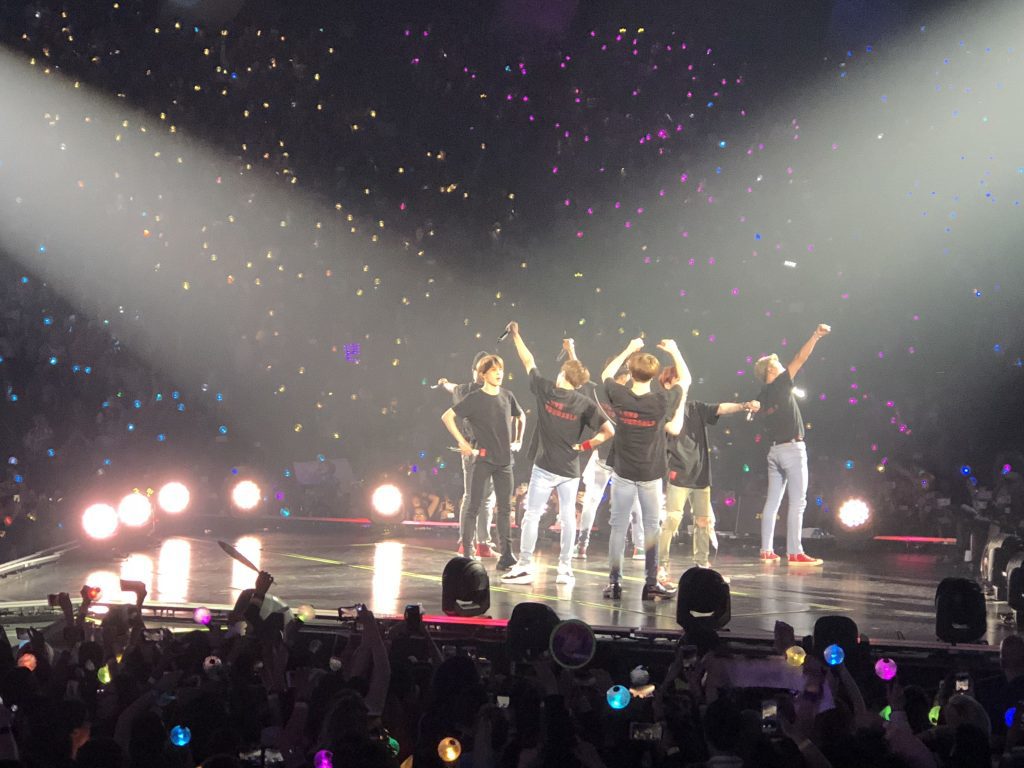 Band BTS on stage waving to fans in audience.