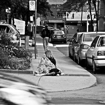 Woman sitting in blanket in middle of sidewalk.