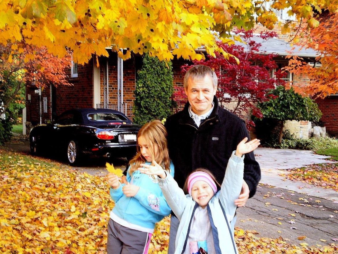 Photo of father and two daughters in leaves outside brick house.