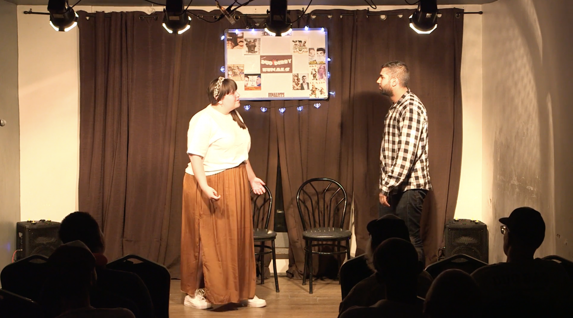Man and woman on stage with black curtain and two chairs.