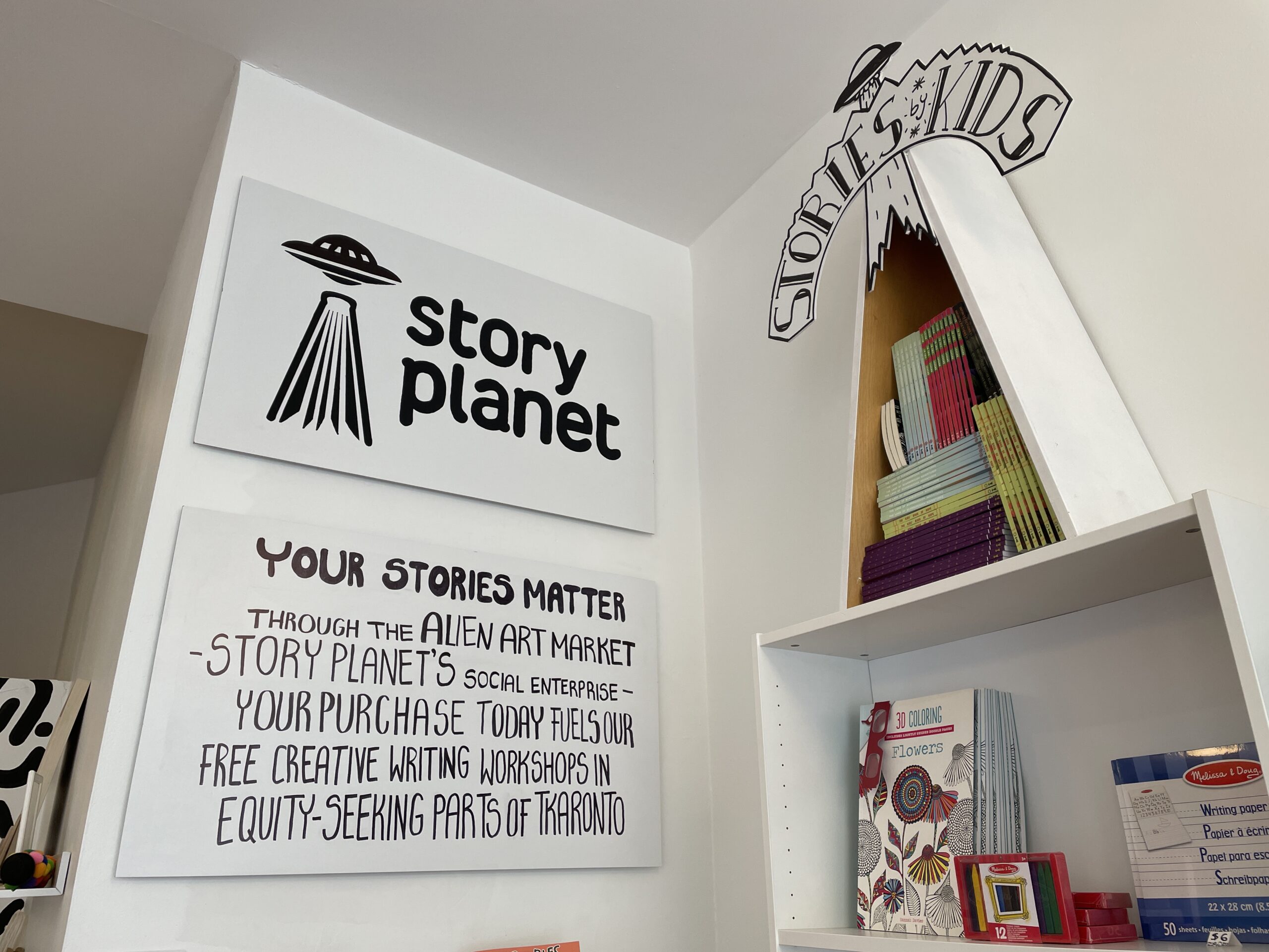 A wall in Alien Art Market with two signs. The first features the logo of Story Planet, which is a UFO next to the words 'Story Planet.' The second sign reads: "Your stories matter. Through the Alien Art Market - Story Planet's social enterprise - your purchase today fuels our free creative writing workshops in equity-seeking parts of Tkaronto."