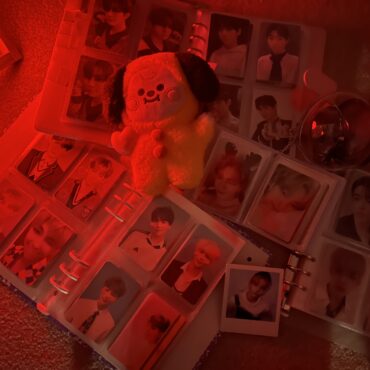 Binders filled with collectible photo cards of different K-Pop idols are strewn across the floor, and lit by a red light. BTS, Seventeen and ENHYPEN are the main groups pictured.