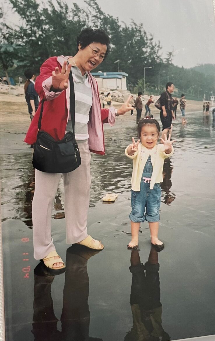 A young child poses with her grandmother cheerfully at a beach.
