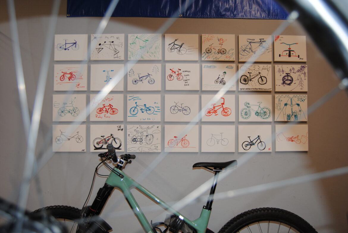 Photograph taken through bike spokes of a light green bike leaned against a photo wall composed of hand drawn illustration of bicycles.