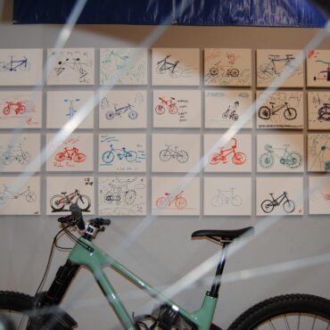 Photograph taken through bike spokes of a light green bike leaned against a photo wall composed of hand drawn illustration of bicycles.