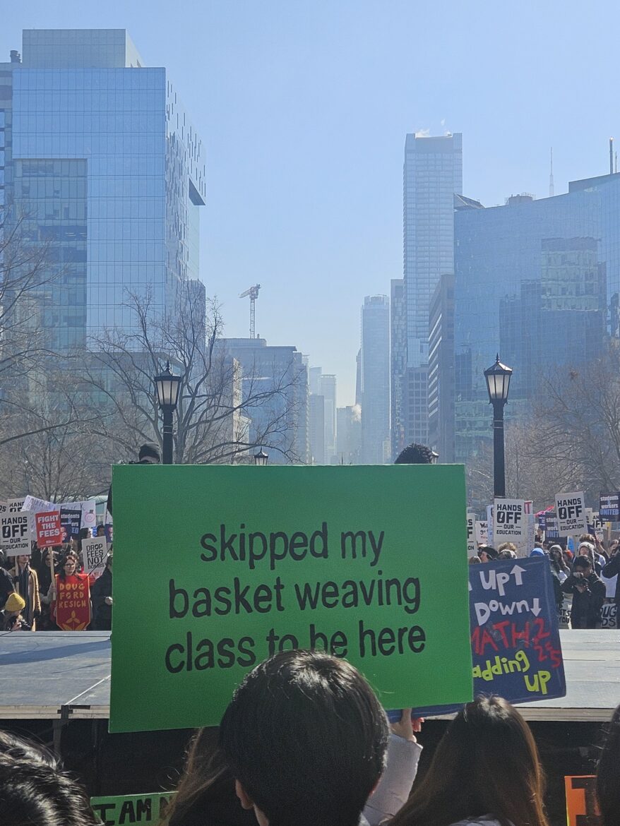 student hold green protest sign that says "skipped my basket weaving class to be here"