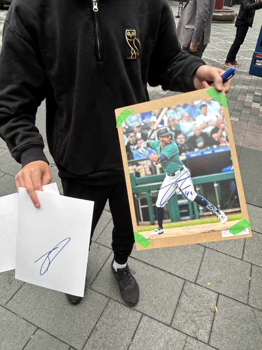 A person wearing a black hoodie holds a signed photo of Seattle Mariners player Julio Rodríguez taped to a board, along with a separate sheet of paper featuring another autograph, standing on a city sidewalk.