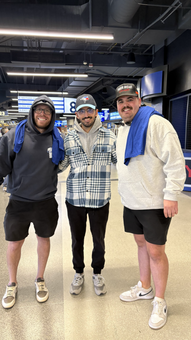 Three men smile and pose together inside a sports arena concourse, wearing casual clothing and hats, with concession stands and section signs visible behind them.
