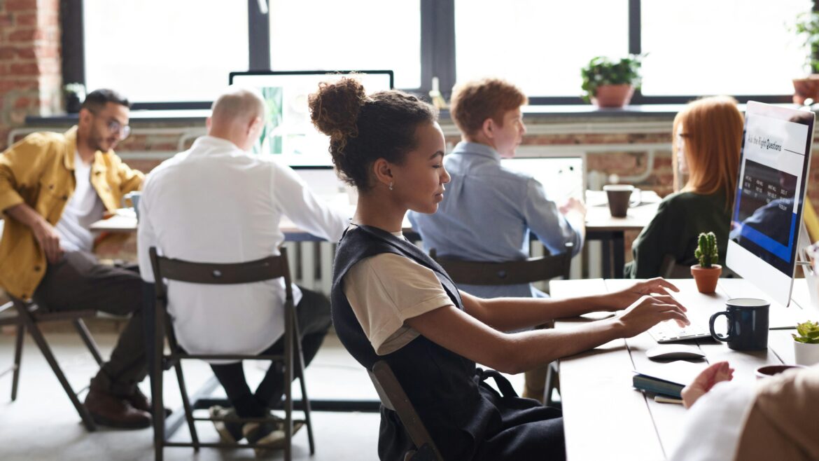 A young woman works at a desktop computer in a modern open office, while several coworkers sit and work at desks in the background.
