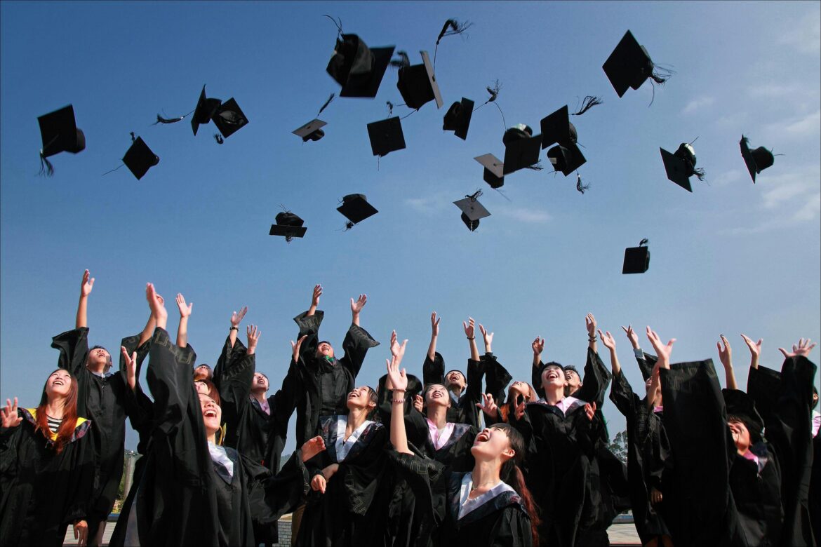 A group of graduates in black caps and gowns joyfully toss their mortarboards into the air under a clear blue sky during a graduation ceremony.