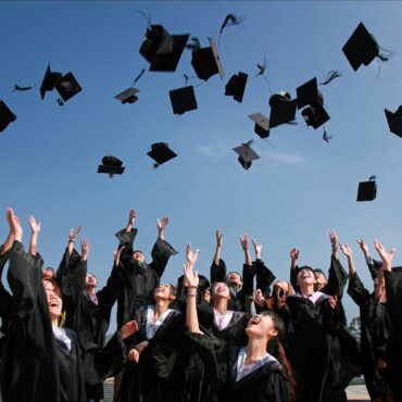 A group of graduates in black caps and gowns joyfully toss their mortarboards into the air under a clear blue sky during a graduation ceremony.
